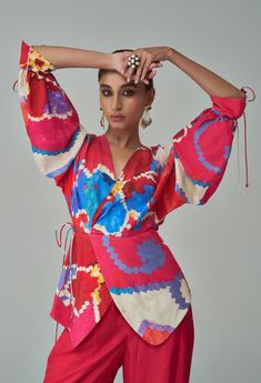 A pink and red ikat print-inspired wrap-style blouse with a tie waist and adjustable tie-up sleeves, paired with high-waist palazzos featuring pockets, creates a vibrant and stylish ensemble. This outfit merges traditional ikat patterns with modern design elements, offering comfort and functionality with a touch of sophistication. Ideal for both casual and semi-formal occasions, it combines bold colors and versatile pieces for a chic and effortless look. Blouse And Palazzo, Print Blouse Design, Ikat Blouse Designs, Ikat Blouse, Pocket Blouse, Palazzo Set, Ikat Print, Indian Fabric, Ikat Pattern