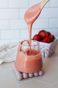 a pink drink being poured into a glass with strawberries in the bowl behind it