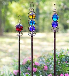 three different colored glass balls on metal poles in front of flowers and trees with pink flowers behind them