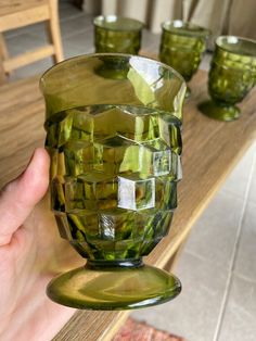 a hand is holding a green glass vase on a table with four glasses in the background