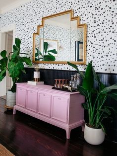 a pink dresser and mirror in a room with black wallpaper, potted plants and a gold framed mirror on the wall