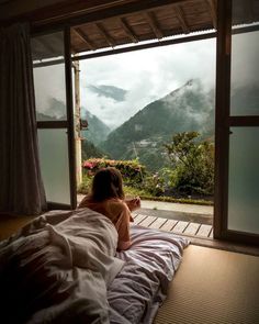 a woman sitting on top of a bed in front of a window next to a lush green hillside