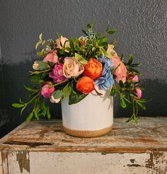 a white vase filled with colorful flowers on top of an old wooden table next to a gray wall