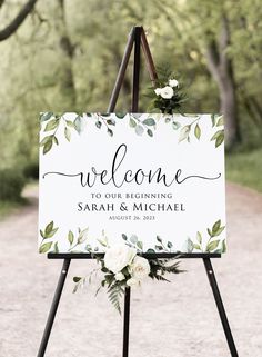 a welcome sign with greenery and white flowers on it is standing in the middle of a dirt road