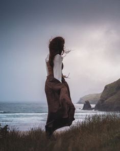 a woman standing on top of a lush green hillside next to the ocean under a cloudy sky