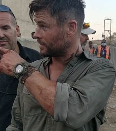 two men standing next to each other in front of some construction workers and one is holding his watch