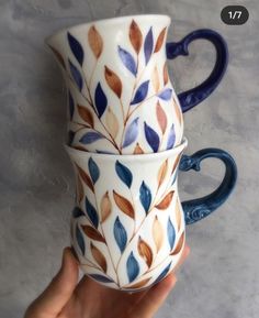 a hand holding two coffee mugs with leaves on the top and bottom, both decorated in blue and brown