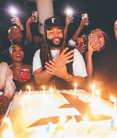 a group of people standing around a birthday cake with candles on it and one person holding his arms up