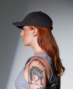 a woman with red hair wearing a baseball cap and sleeve tattoos on her arm, looking off to the side