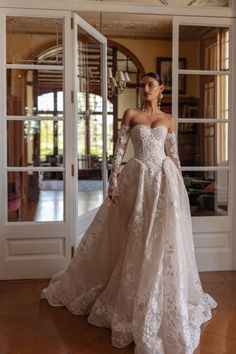 a woman standing in front of a doorway wearing a wedding dress with long sleeves and an off the shoulder design