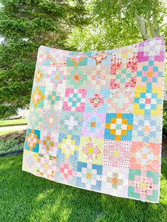 a colorful quilt hanging from a tree on the grass in front of some green trees