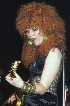 a woman with long red hair holding a guitar in her right hand and wearing green earrings