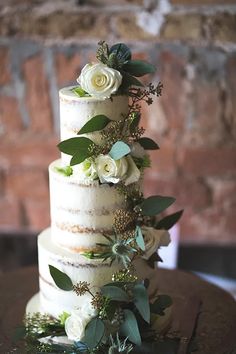 a three tiered cake with white flowers and greenery on the top is displayed in front of a brick wall