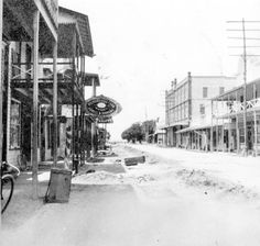 an old black and white photo of a town with snow on the ground in front of buildings