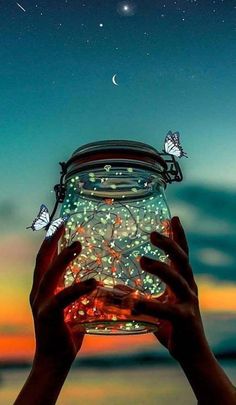 a person holding up a jar with butterflies on it at the beach under a night sky