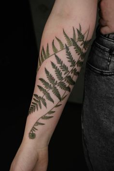 a fern leaf tattoo on the left forearm and arm, with green leaves growing out of it