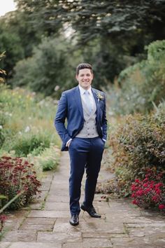 a man in a blue suit and tie standing on a brick path surrounded by flowers