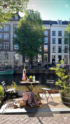 there is a table and chairs on the side of the river with buildings in the background