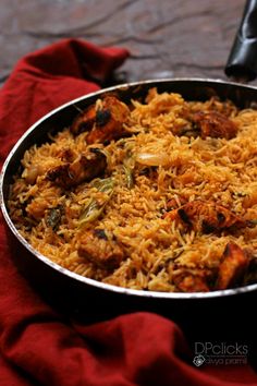 a pan filled with rice and meat on top of a red cloth next to a knife