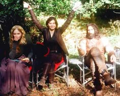 three people sitting on chairs in the woods with one holding up his arms and smiling at the camera