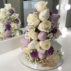 a white and purple wedding cake with flowers on the table in front of a mirror