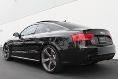 the rear end of a black car parked in front of a white wall and building