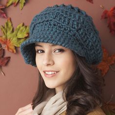 a woman wearing a blue crocheted hat and scarf with autumn leaves on the wall behind her