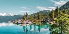 a lake surrounded by trees and mountains with blue water in the foreground on a sunny day