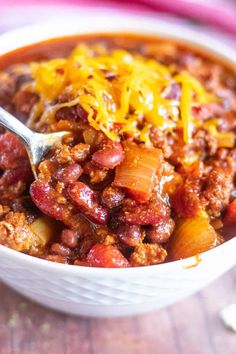 a white bowl filled with chili and beans on top of a wooden table next to a spoon