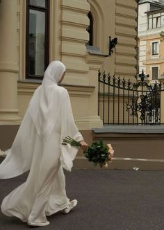 a woman dressed in white walking down the street with flowers on her hand and wearing a veil over her head