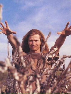 a man with long hair and hands up in the air, surrounded by bushes on a sunny day