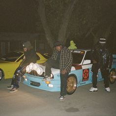 two men sitting on the hood of a race car at night with other people standing around