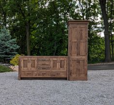 a large wooden bed sitting in the middle of a gravel field next to trees and bushes