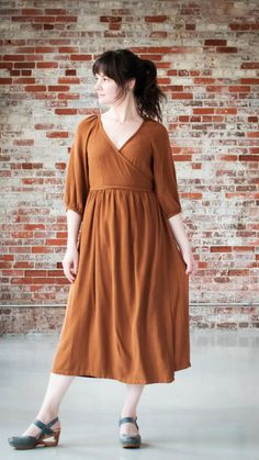 a woman standing in front of a brick wall wearing a brown dress and black sandals