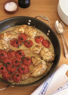 a pan filled with chicken and tomatoes on top of a wooden table next to utensils