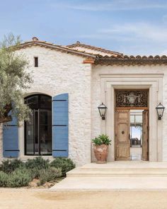 the front entrance to a white stucco house with blue shutters and an olive tree