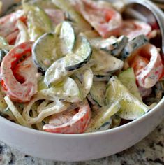 a white bowl filled with pasta salad on top of a marble counter topped with tomatoes and cucumbers