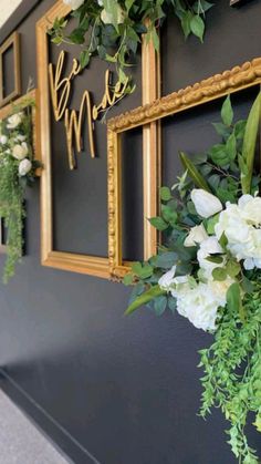 flowers and greenery are hanging on the wall in front of a gold framed sign