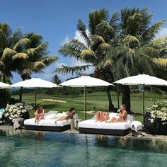 three women lounging on lounge chairs next to a pool with umbrellas over them