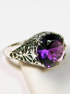 This elegant ring features a stunning Art Nouveau design in sterling silver. The focal point is a 3.5ct round, full cut Amethyst in a vibrant purple hue. You can choose between a Vs to VVS Simulated or Natural Amethyst, with the color of the natural stone being close to the one shown in the photo (Choose Gemstone above). The Amethyst measures 10mm (3/8th of an inch) in diameter and adds a touch of glamour to the delicate filigree setting. The band is marked 925 for sterling silver. The filigree Purple Promise Ring With Intricate Design, Purple Intricate Design Promise Ring, Purple Hallmarked Crystal Wedding Ring, Classic Purple Crystal Round Ring, Classic Round Purple Crystal Ring, Classic Purple Round Crystal Ring, Classic Purple Crystal Ring, Exquisite Round Amethyst Ring With Prong Setting, Amethyst Ring With Intricate Design