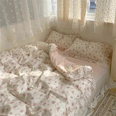 an unmade bed sitting in front of a window with white curtains and pink flowers on it