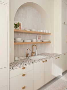 a kitchen with white cabinets and gold faucets on the counter top, along with shelves filled with dishes