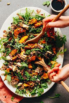 two hands holding wooden utensils over a plate of salad with peaches and greens