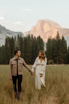 a man and woman holding hands while walking through tall grass in front of a mountain