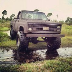 an old pick up truck is parked in the mud near some grass and water on a sunny day