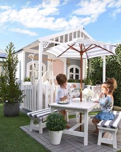 two children sitting at a table with an umbrella in the back ground, and another child standing next to it