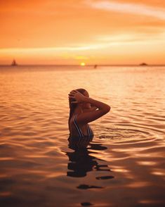 a woman sitting in the water with her arms behind her head, looking at the sunset