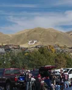 a group of people standing around parked cars in a parking lot with mountains in the background