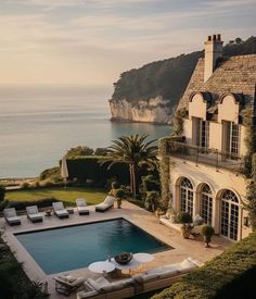 an outdoor swimming pool next to a large house near the ocean with lounge chairs around it