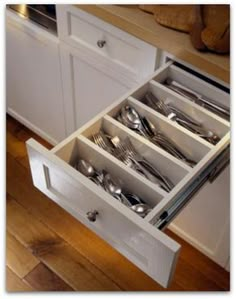 an open drawer with silverware in it on top of a wooden floor next to white cabinets